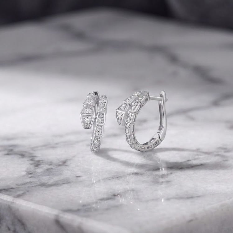 Two silver hoop earrings with geometric and diamond-like patterns are displayed on a gray marble surface, reflecting light from above. These 14k White Gold Snake Diamond Hoop Earrings 1/3CTW feature a luxurious modern design, adding elegance to any look.