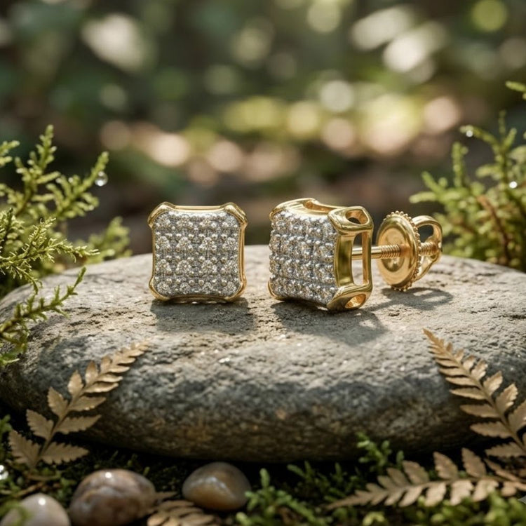 A pair of 10K Gold Diamond Square Earrings (1/10 CTTW) rest on a smooth stone, surrounded by green ferns and small pebbles, outdoors in soft natural light.