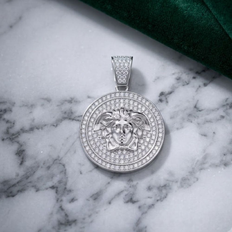 A round Moissanite Medusa Egyptian Charm Pendant, crafted in sterling silver and adorned with sparkling stones, featuring a striking Medusa head design, rests on a white marble surface with a green textured fabric in the background.