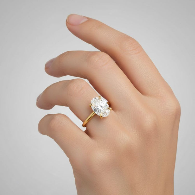 A hand with neatly manicured nails wears a 1.72ct oval-cut lab diamond engagement ring in 14k solid yellow gold on the ring finger against a plain gray background.