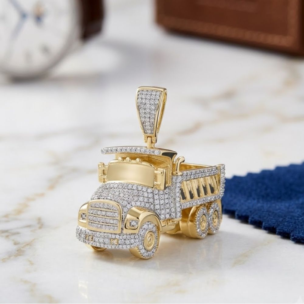 The 7/8 CT Diamond Truck Charm in 10K Yellow Gold is shaped like a dump truck with natural diamonds, displayed on marble with a blurred watch and blue cloth in the background.