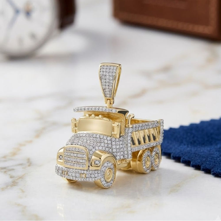 The 7/8 CT Diamond Truck Charm in 10K Yellow Gold is shaped like a dump truck with natural diamonds, displayed on marble with a blurred watch and blue cloth in the background.