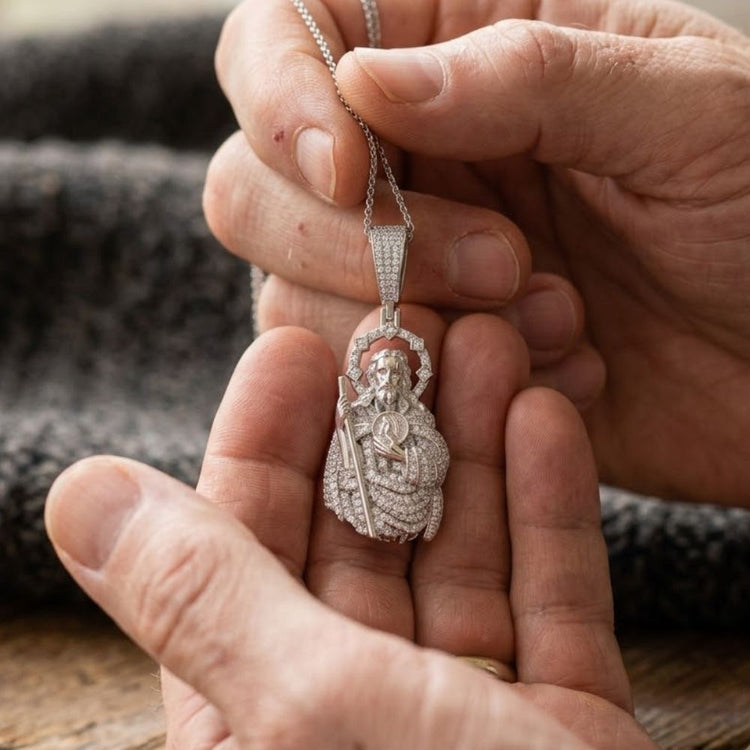 A person holds the Classic Moissanite St. Jude Charm Pendant—a silver-toned accessory crafted from hypoallergenic 925 sterling silver, shaped like a crowned figure with a sword and adorned with sparkling stones—over textured gray fabric.