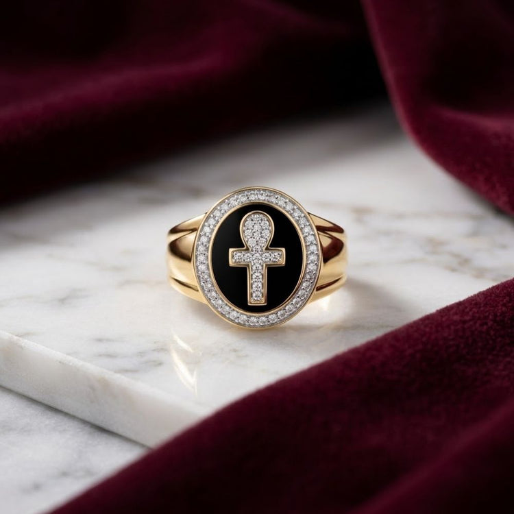 A captivating 10k Yellow Gold Diamond Ankh Cross Ring 1/6 Cttw with a black circular face showcases a diamond-adorned ankh symbol, elegantly displayed on a white marble surface and accented by burgundy velvet fabric.