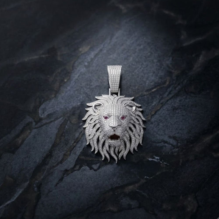 The Silver Moissanite Enameled Lion Head Charm Pendant, encrusted with diamond accents and featuring vivid red gemstone eyes, rests on a dark, textured stone surface.
