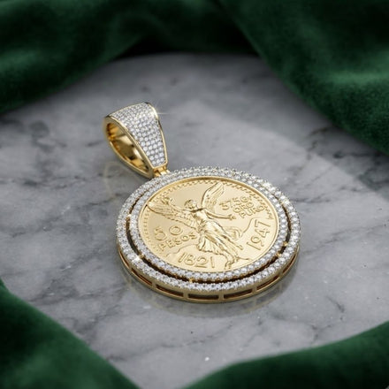 The Diamond Centenario Spinning Pendant in 14K yellow gold (1.875CT) features a detailed design, diamond-studded border, and 50 Pesos 1821-1947 coin with a winged figure, displayed on gray marble with green fabric.