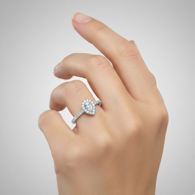 A hand with neatly manicured nails wears the Elegant Pear-Shaped Lab Diamond Halo Ring, featuring a dazzling pear-shaped diamond cluster encircled by smaller diamonds, set against a plain light gray background.