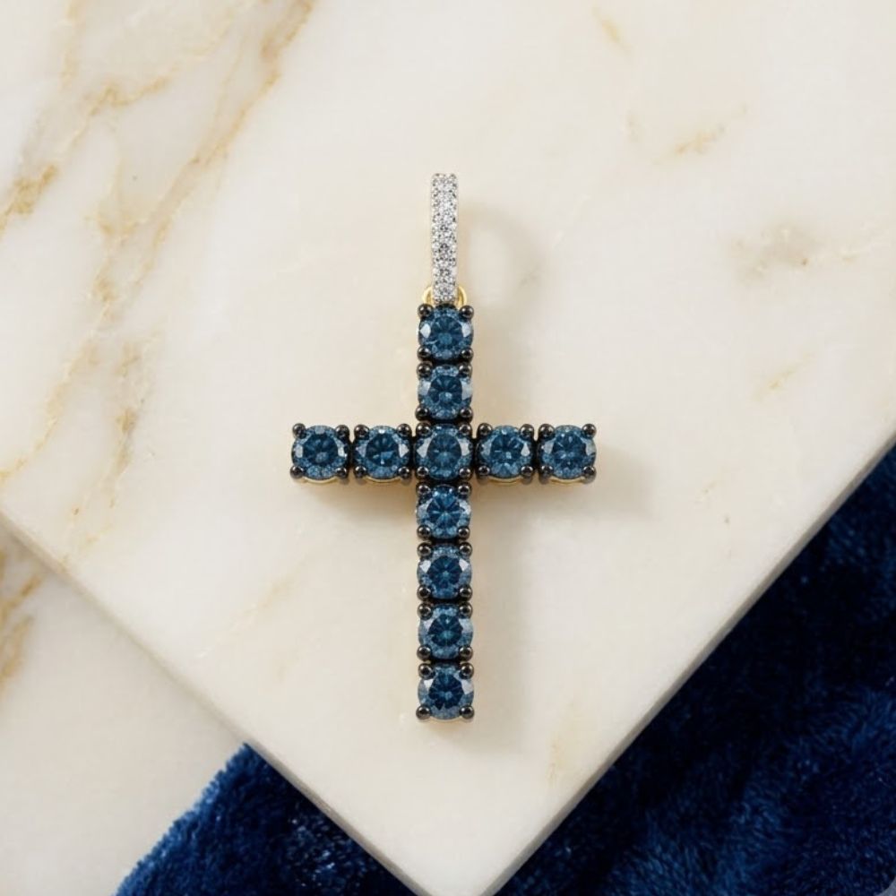 The 14K Yellow Gold Blue Diamond Cross Pendant, featuring round blue diamonds and a diamond-studded vertical bail, rests on white marble beside deep blue velvet fabric.