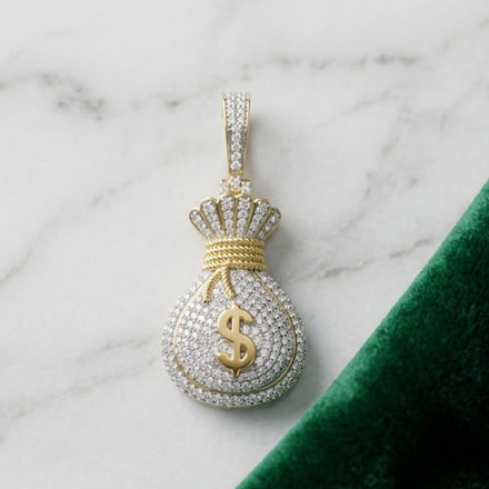 The Diamond Money Bag Charm 7/8 CTW in 10K yellow gold, shaped with a dollar sign, is shown on white marble beside green velvet—an eye-catching men's hip hop jewelry piece.