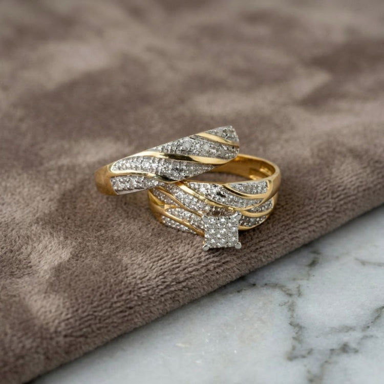 Two gold rings featuring intertwined silver bands and sparkling diamonds rest gently on soft brown fabric beside a marble surface—an elegant display of the 1/3 CTW 10k Yellow Gold Wedding Trio Set, ideal for those seeking an exquisite engagement ring and coordinating wedding bands.