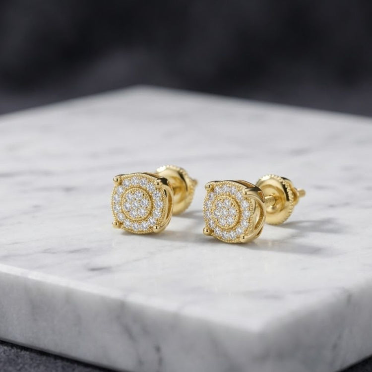 A pair of 925 Sterling Silver Yellow-Tone Moissanite Studs rests on a white marble slab with a gray, blurred background.