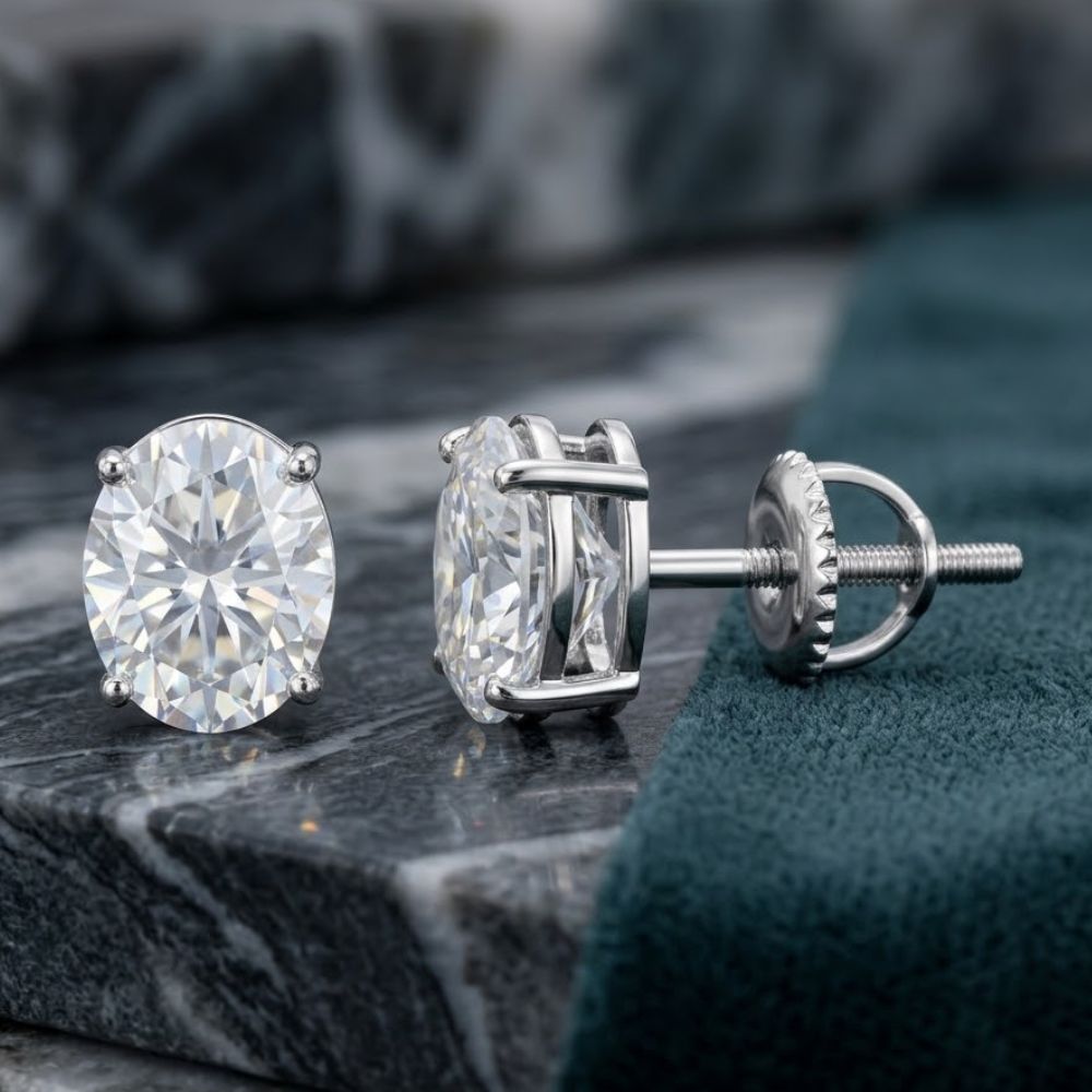 A pair of 925 Sterling Silver Oval Moissanite Stud Earrings with prong settings and screw-back posts, displayed on a gray marble surface beside dark green fabric.