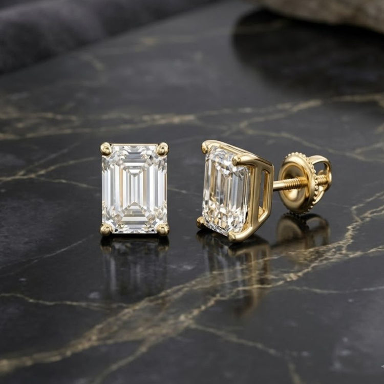 Two gold stud earrings with rectangular, emerald-cut clear gemstones are displayed on a dark, marble-patterned surface, showcasing the timeless elegance of the 14K Yellow Gold 1/4 CTW Emerald Solitaire Earrings.