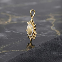 The 10k Yellow Gold Diamond Gift Pendant 1/10 Cttw, featuring a gold sunflower shape with a central oval and a ring of sparkling diamonds, is displayed on a black marble surface beside a textured dark cloth.