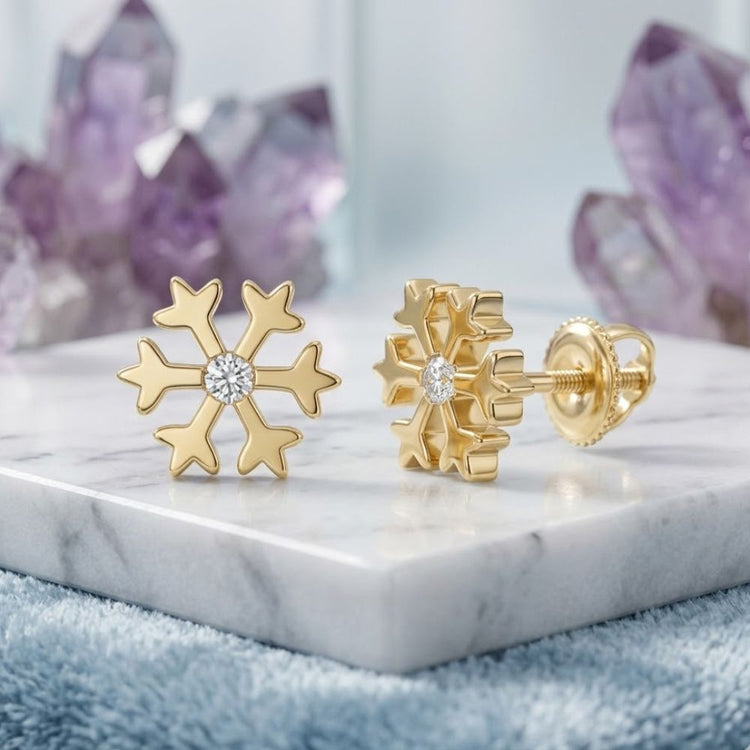 The 10k Yellow Gold Diamond Snowflake Earrings 1/20 CTTW are displayed on a white marble surface with purple crystals in the background.