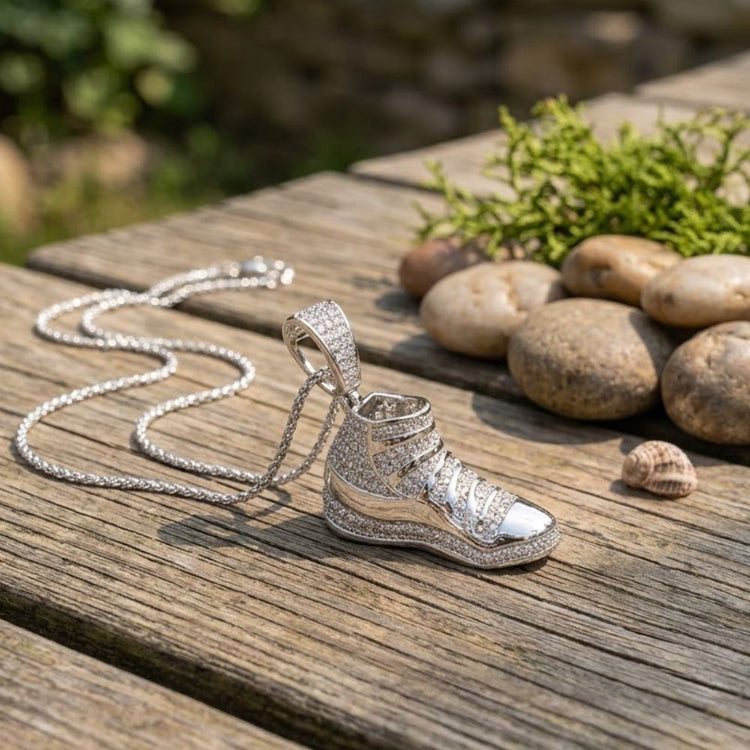 The Sterling Silver Diamond Shoe Pendant 1/2 CTW, featuring a sneaker-shaped pendant, rests on a wooden surface outdoors near smooth stones, green moss, and a small seashell—this striking piece adds sparkling elegance and unique charm to any look, making it the perfect statement jewelry.