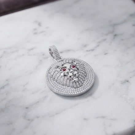 The Silver Ice Out Lion Head Charm Pendant, featuring a round shape and encrusted with sparkling VVS moissanite stones along with two vivid red gemstone eyes, rests elegantly on a white marble surface. Expertly crafted from hypoallergenic sterling silver, this dazzling piece offers both luxurious style and comfortable wear.