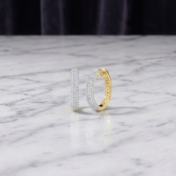 A pair of 3/4CTW Diamond Hoop Earrings in 10k yellow gold, adorned with small natural diamonds, displayed upright on a white marble surface with a dark, blurred background.