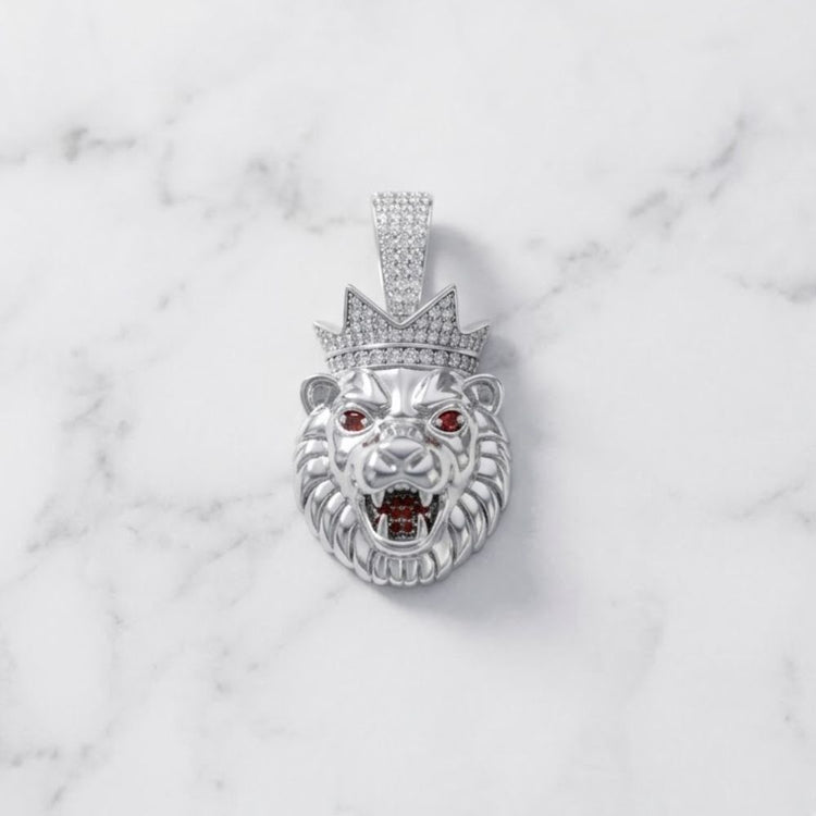 The King Lion Head Charm Pendant - Silver Moissanite is a striking sterling silver piece, showcasing bold red gemstone eyes and an open mouth beneath a crown encrusted with sparkling moissanite stones. This intricately detailed pendant rests elegantly on a white marble surface.
