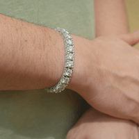 A close-up of a wrist wearing the Fully Iced Out Moissanite Tennis Bracelet in 925 sterling silver, with round, clear gemstones sparkling. The person rests their other hand on their wrist and wears a light green shirt.