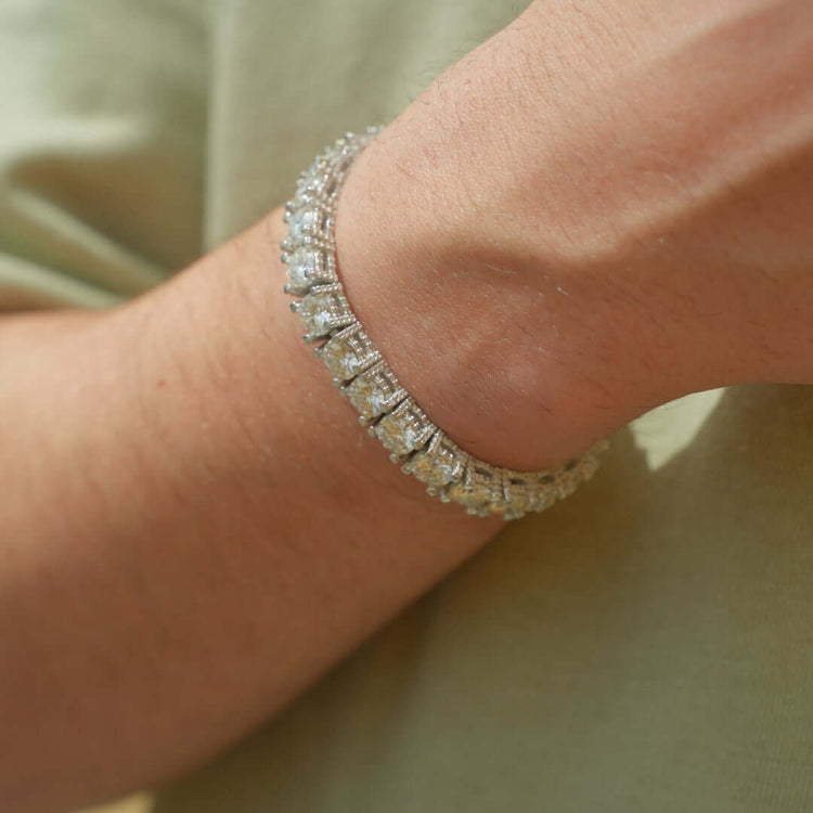 A close-up of a wrist wearing the Fully Iced Out Moissanite Tennis Bracelet, sparkling with rectangular and round stones set in 925 sterling silver, against a light green shirt.