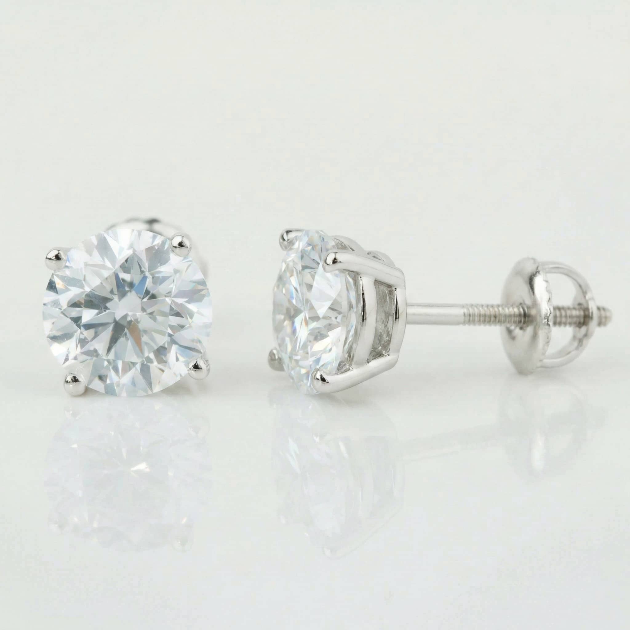 The 14k Classic Round Lab Diamond Four Prong Stud Earrings (F/G VS+) feature round lab diamonds in 14K white gold prongs with secure screw-back posts, showcased on a sleek white reflective surface.