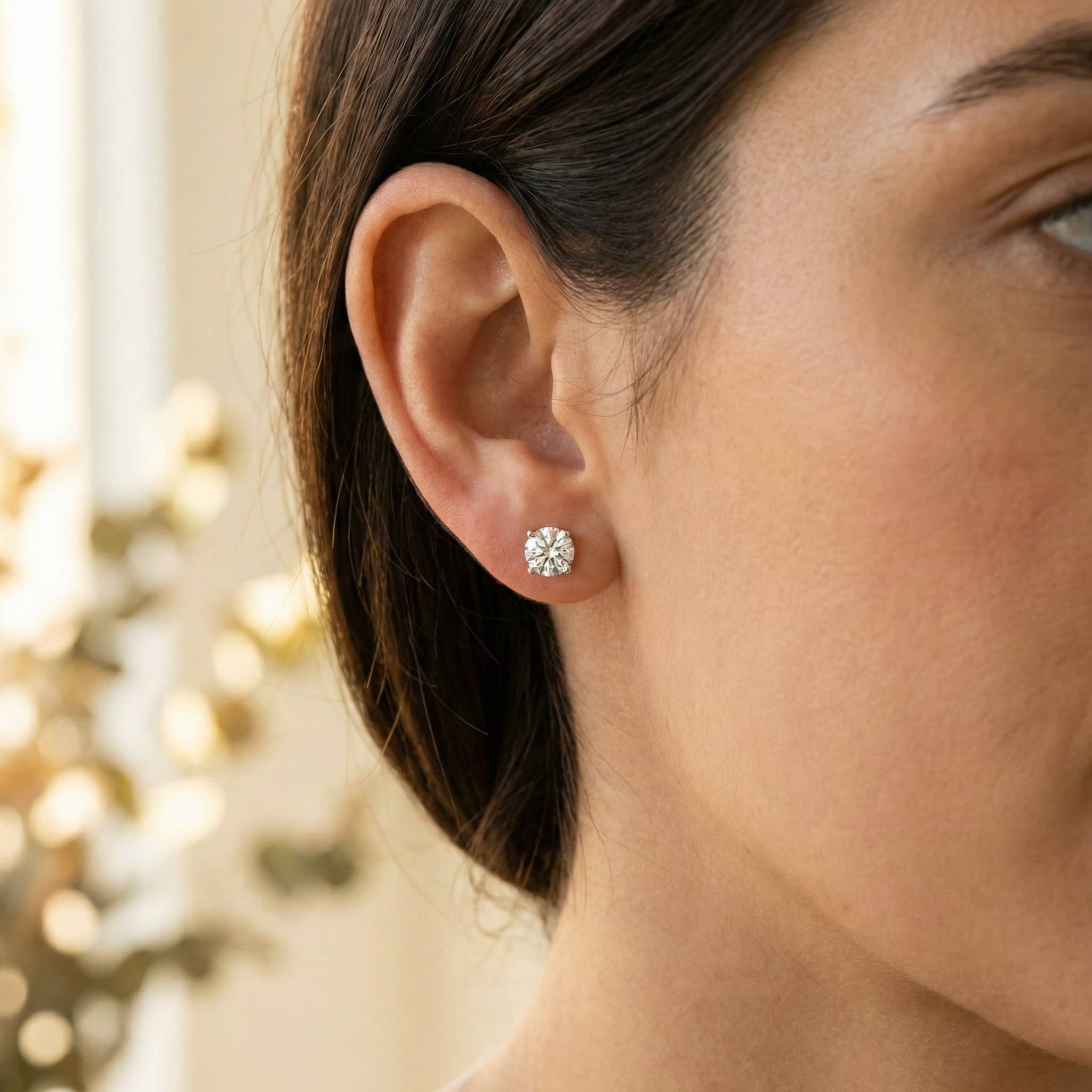 Close-up of a woman's ear wearing 14k Classic Round Lab Diamond Four Prong Stud Earrings (F/G VS+). The softly blurred background is lit with warm, natural light.