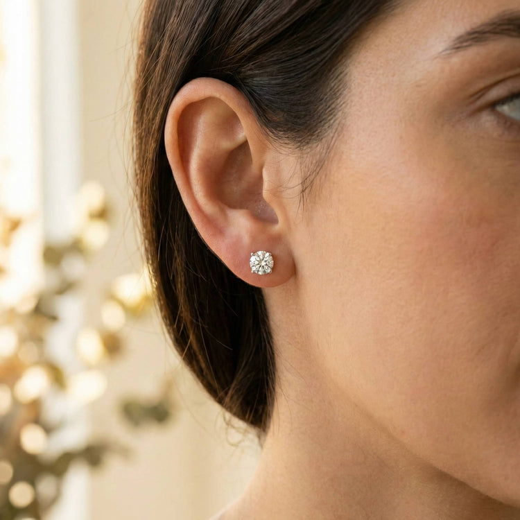 Close-up of a woman's ear wearing 14k Classic Round Lab Diamond Four Prong Stud Earrings (F/G VS+). The softly blurred background is lit with warm, natural light.