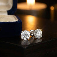 A pair of 14k Classic Round Lab Diamond Four Prong Stud Earrings (F/G VS+) rest on a dark surface, sparkling under warm lighting, with a velvet jewelry box and a softly glowing candle in the background.