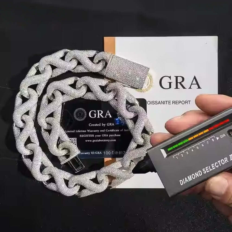 A hand uses a diamond tester on the Iced Out Silver Moissanite Cuban Link Bracelet - 20mm. Behind the sterling silver bracelet are a GRA certificate, warranty card, and orange envelope. The tester’s lights glow as it checks the jewelry.