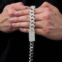 A person holds a Moissanite Cuban Link Chain (12mm-15mm, 925 Sterling Silver) encrusted with sparkling stones in both hands against a black background.