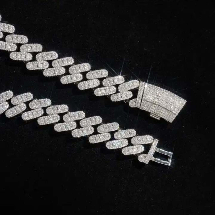 The Moissanite Cuban Link Chain (12mm-15mm) in 925 Sterling Silver sparkles with an iced-out shine, its brilliant clasp gleaming against a black background.