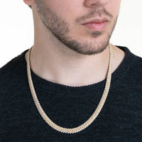 A man with short facial hair wears the Moissanite Cuban Link Chain 8mm-23mm in solid yellow gold over a textured black sweater. The image focuses on his neck, jawline, and lips against a plain background.