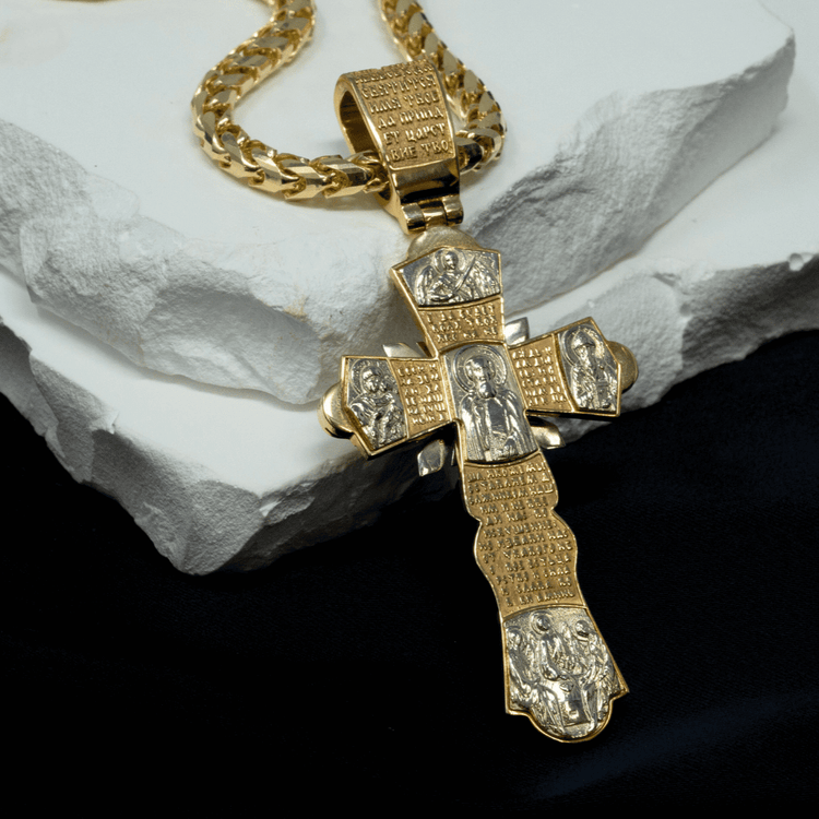 The Orthodox Cross Pendant - 14K Gold features engraved religious figures and inscriptions on an ornate cross, hanging from a gold chain. It is displayed on a white stone slab with a black cloth background.