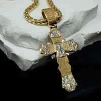 The Orthodox Cross Pendant - 14K Gold features engraved religious figures and inscriptions on an ornate cross, hanging from a gold chain. It is displayed on a white stone slab with a black cloth background.