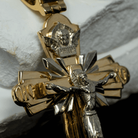 A close-up of the Orthodox Cross Pendant - 14K Gold shows Jesus on the cross with fine details and a small angel above Him, representing Orthodox Christianity. The pendant lies on a white textured background.