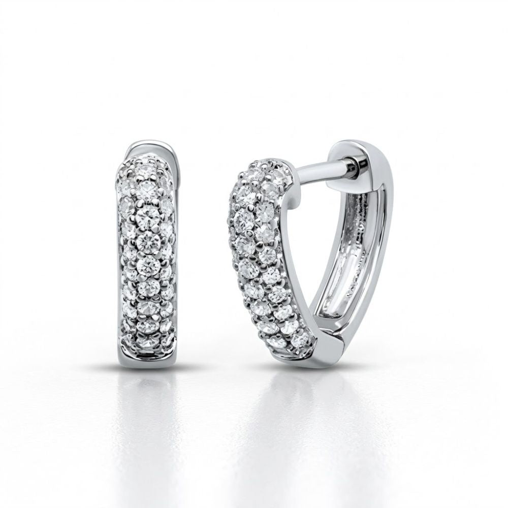 The 10K White Gold Diamond Heart Huggie Hoop Earrings are shown on a white background, with one earring upright and the other on its side to display the latch-back closure. Both feature small round diamonds for added sparkle.