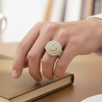 A hand rests on a closed book, wearing the Layered Round Diamond Ring – 2.33 CT Gold Design, encrusted with rows of sparkling diamonds. The softly blurred background creates an elegant indoor setting—ideal for showcasing gold men’s rings.