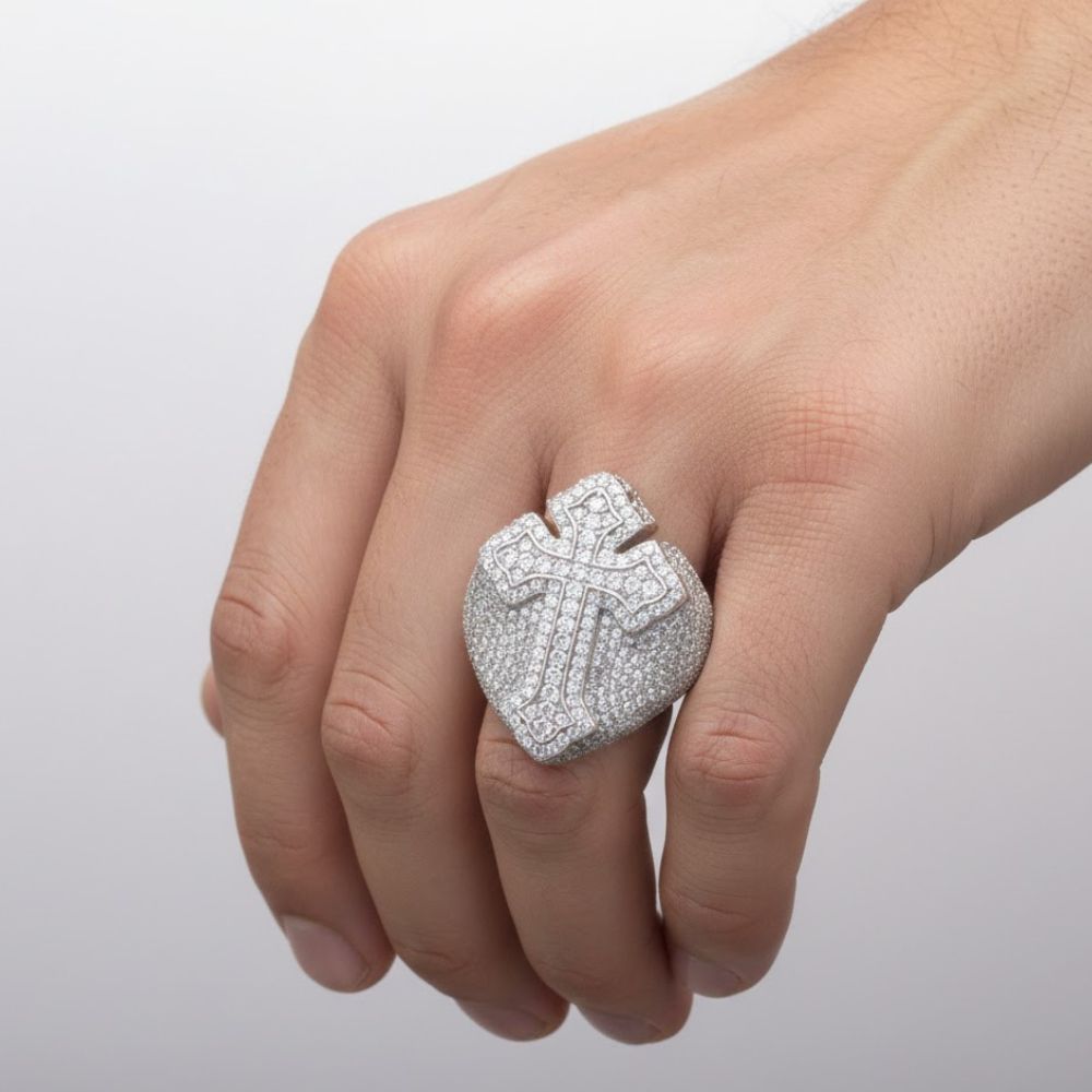 A hand wears the Moissanite Cross Religious Ring, a large silver shield-shaped band with a sparkling cross design encrusted with diamonds. Set against a plain light background, this elegant faith jewelry piece stands out beautifully.