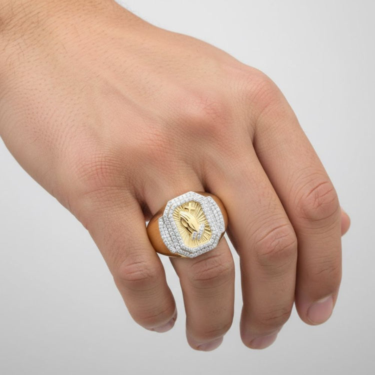 A hand displays the 1/2CTW Diamond Praying Hands Cross Men’s Ring in 10k gold, featuring an engraved praying hands cross and a border of small clear stones, set against a plain, light-colored background.