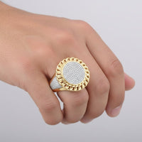 A hand displays the 10K Yellow Gold Diamond Cuban Link Ring 1/2CT, featuring a round diamond face with a twisted gold border and sparkling accents, shown against a simple light background.