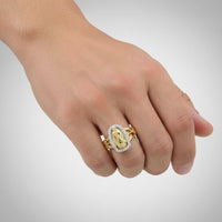 A hand wears the 1/5CTW 14k Gold Virgin Mary Ladies Ring, featuring an engraved central image, set against a plain light gray background.
