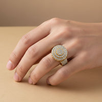 A hand rests on a beige surface, showcasing the Layered Round Diamond Ring – 2.33 CT Gold Design, a bold men's gold ring featuring multiple rows of sparkling white diamonds arranged in concentric circles.