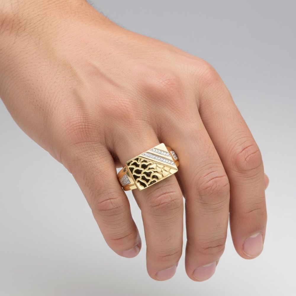 A hand displays the 10k Yellow Gold Diamond Nugget Ring 1/20 Cttw, featuring a rectangular face with a bold black pattern and diamond accents, set against a plain light gray background.