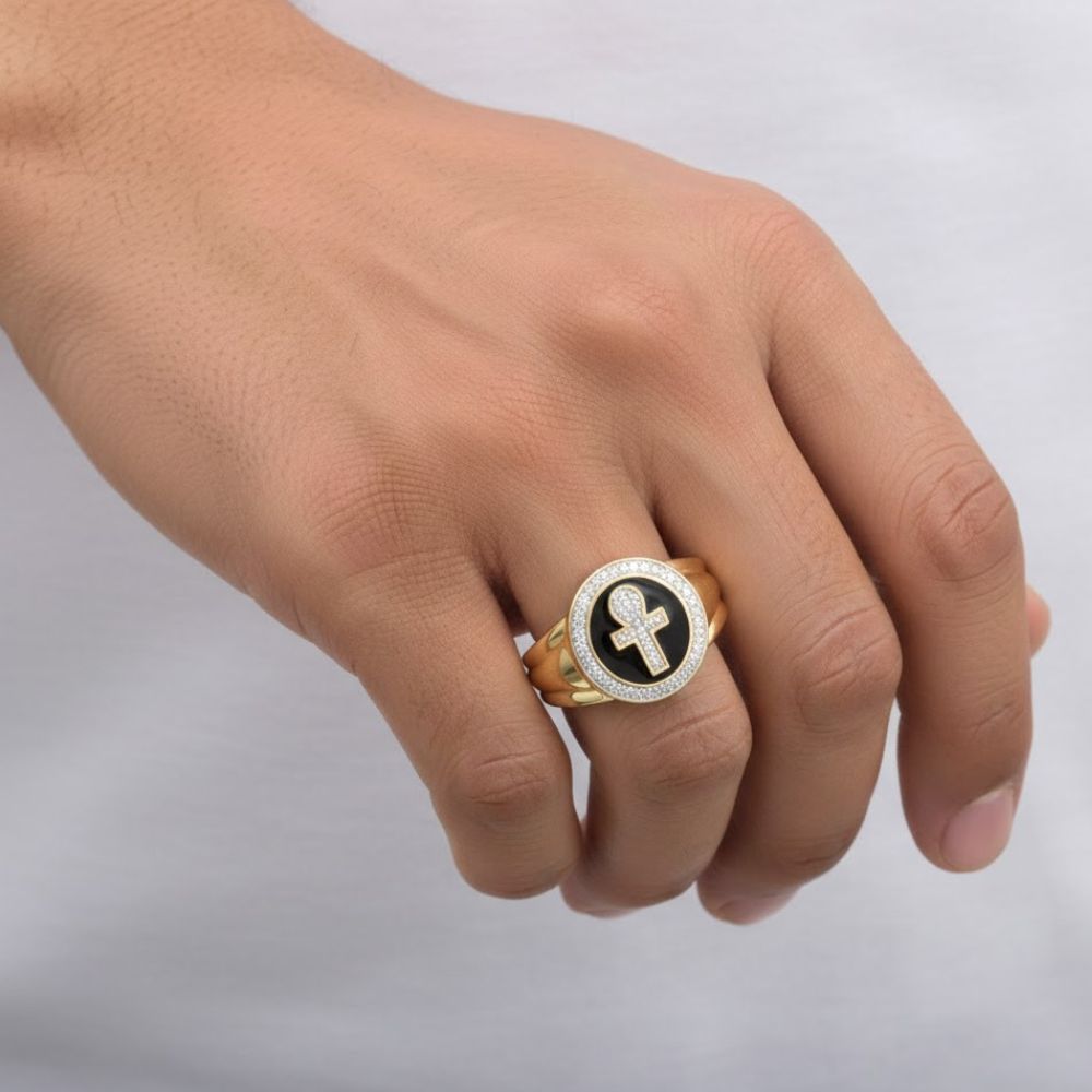 A hand wears the 10k Yellow Gold Diamond Ankh Cross Ring 1/6 Cttw, featuring a black circular face with a central cross and clear stones. A white shirt highlights this striking piece of faith jewelry.