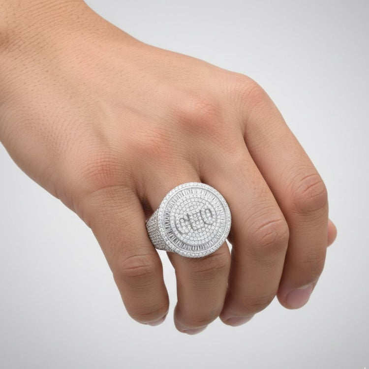 A hand wears the Baguette Moissanite CEO Round Head Fashion Ring, featuring a large round silver setting with diamond accents and "CEO" in the center, shown against a plain light background.