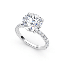 The Round Cut Lab Diamond Engagement Ring with Pave Band features a large round center stone in a four-prong setting and a pavé band with smaller diamonds, available in white gold or platinum.