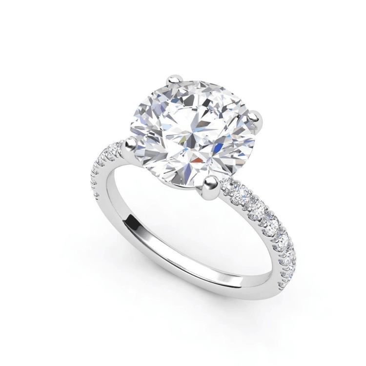The Round Cut Lab Diamond Engagement Ring with Pave Band features a large round center stone in a four-prong setting and a pavé band with smaller diamonds, available in white gold or platinum.