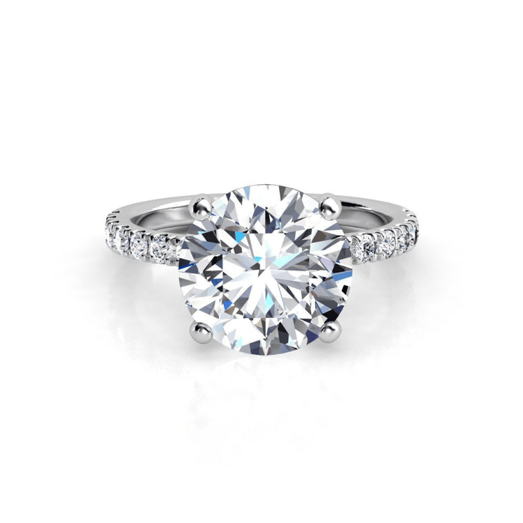 The Round Cut Lab Diamond Engagement Ring with Pave Band features a silver band, a large round lab diamond in a four-prong setting, and pavé-set smaller diamonds along the band.