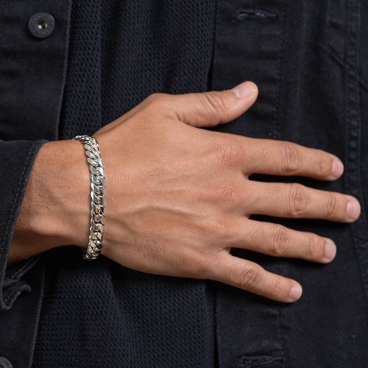 A close-up of a person's hand on their chest, wearing a 10mm Handmade Miami Cuban Link Bracelet - 950 Silver and a black textured shirt or jacket.